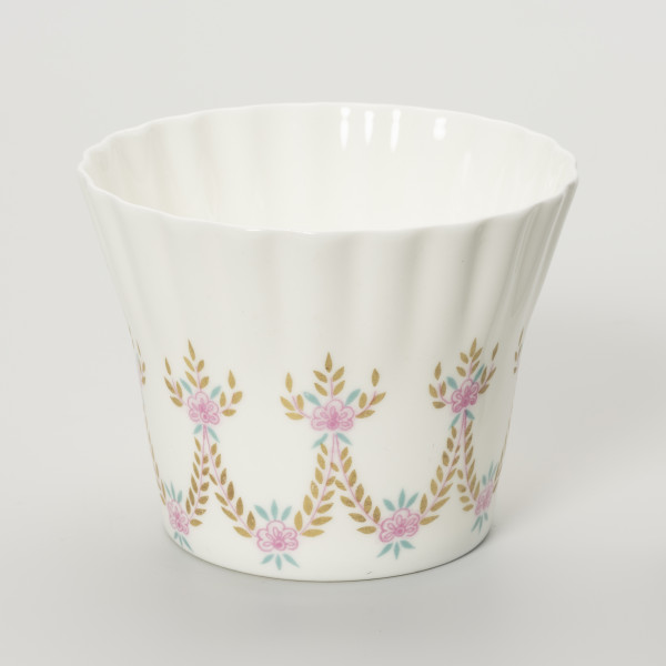 Filiżanka - ujęcie z drugiego boku; Duża filiżanka z białej porcelany o lekko dzwonowatej formie, z płaskim dnem i karbowaną górną częścią czarki. Dekorowana malowanymi festonami z wici roślinno-kwiatowej w barwach różu, zieleni i starego złota.