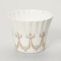 Filiżanka - ujęcie z drugiego boku; Duża filiżanka z białej porcelany o lekko dzwonowatej formie, z płaskim dnem i karbowaną górną częścią czarki. Dekorowana malowanymi festonami z wici roślinno-kwiatowej w barwach różu, zieleni i starego złota.