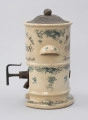 Pojemnik na musztardę - ujęcie z prawego boku; Cylindryczny, porcelitowy pojemnik w kolorze kremowym na wysokiej podstawie z dozownikiem i pokrywą. Stożkowata blaszana pokrywka z wypłaszczonym rondem i okrągłym uchwytem, fakturowana cyzelowanymi wydłużonymi owalami. Pojemnik cylindryczny. Na wprost wklęsły od spodu, zaokrąglony uchwyt. Na górnej części po bokach fragmenty czarnego, ozdobnego napisu w trzech wierszach, z których pierwszy zapisany po łuku. Porcelitowa podstawa pojemnika w formie pustego cylindra bez dna z wywiniętą na zewnątrz dolną krawędzią markującą stopę oraz plastycznym pierścieniem nieco powyżej. Z lewej strony wystaje metalowy wąski dozownik. Dekorowany granatową kalkomanią: na podstawie, brzegach obu porcelitowych części wzór rocailliowy w dookolnych pasmach, na uchwytach oraz punktowo na podstawie i pojemniku gałązki róż.