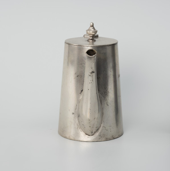 Dzbanuszek z zastawy Konzerthaus Stettin - ujęcie z lewego boku; Dzbanuszek o prostej, zgeometryzowanej formie, bez wykształconej stopy. Korpus cylindryczny, zwężający się ku górze, zamykany płaską, okrągłą pokrywą z gałką w centrum. Dziubek osadzony w dolnej części korpusu, zwężający się ku górze, łagodnie wygięty, sięga wysokości pokrywy. Pokrywa - krążek z blachy.