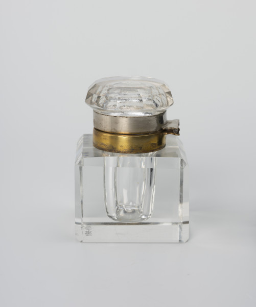 Kałamarz - ujęcie z tyłu; Kałamarz ze szkła kryształowego z metalowym kołnierzem oprawy ujmującej pokrywę. Pojemnik w formie graniastosłupa z szeroko sfazowanymi krawędziami i wklęsłym cylindrycznym wnętrzem o średnicy 2.25 cm przeznaczonym na atrament. Ponad otworem kołnierz ujęty w metalową oprawę z posrebrzanego mosiądzu z zawiasem trzymającym pokrywę. Pokrywa ze szkła lanego z zagłębionym wnętrzem i kołnierzem analogicznym jak w części głównej, ujętym w metalową oprawę z posrebrzanego mosiądzu, która ujmuje kołnierz pierścieniem z otokiem, do którego przymocowana jest górna część zawiasa. Górna część pokrywy okrągła z podwójnym szlifem tworzącym 12-ścienne łagodne zwężenie. Część korpusowa dzięki kryształowej przejrzystości tworzy zwierciadlane odbicia, powielające efekt szlifowanych krawędzi naczynia.