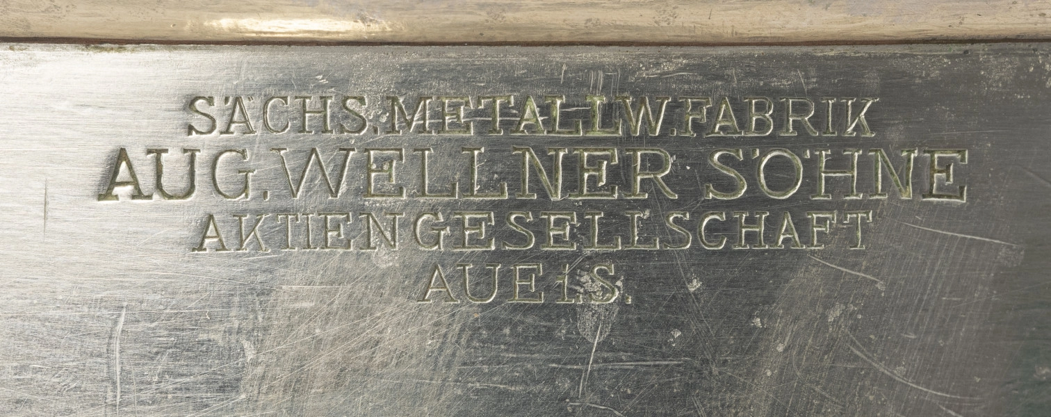 Kubełek do chłodzenia szampana szczecińskiego Knzerthaus - detal; W centrum cylindrycznej części czary napis grawerowany pisanką: SÄCHS. METALLW.FABRIK AUG. WELLNER SÖHNE AKTIENGESELLSCHAFT AUE i.S.