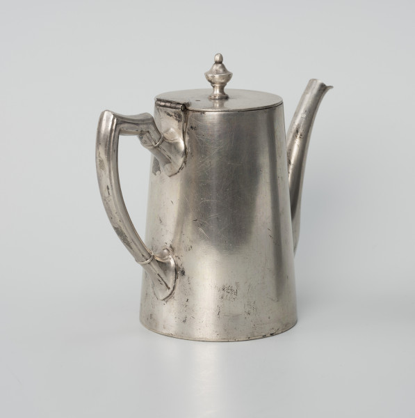 Dzbanuszek z zastawy Konzerthaus Stettin - ujęcie ze skosu z tyłu; Dzbanuszek o prostej, zgeometryzowanej formie, bez wykształconej stopy. Korpus cylindryczny, zwężający się ku górze, zamykany płaską, okrągłą pokrywą z gałką w centrum. Dziubek osadzony w dolnej części korpusu, zwężający się ku górze, łagodnie wygięty, sięga wysokości pokrywy. Ucho o czworobocznym przekroju z profilem (rowkiem) na krawędziach, górą wyniesione do wysokości pokrywy, a następnie załamane, poprowadzone niemal poziomo i powtórnie załamane, a dalej delikatnie wygięte i łukiem wprowadzone do ścianki korpusu. Górą i dołem powyżej wygięcia przewiązka. Pokrywa - krążek z blachy na zawiasie powyżej ucha; uchwyt umocowany centralnie, w formie kielichowej gałki zwieńczonej kulką.