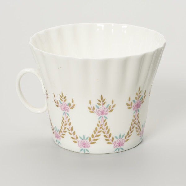 Filiżanka - ujęcie z tyłu; Duża filiżanka z białej porcelany o lekko dzwonowatej formie, z płaskim dnem i karbowaną górną częścią czarki. Z lewej strony proste, owalne doklejone pośrodku wysokości ucho. Filiżanka dekorowana malowanymi festonami z wici roślinno-kwiatowej w barwach różu, zieleni i starego złota.