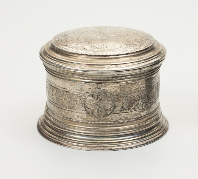 Puszka liturgiczna na komunikanty - ujęcie z drugiego boku; Okrągła puszka liturgiczna na komunikanty, wykonana z pozłacanego srebra. Wieczko okrągłe, z podwójnym, niezdobionym kołnierzem. Na wieczku ryty motyw roślinny. Brzeg wieczka ze srebrnymi granulkami. Na ściance szeroki, okrągły pas ze zdobieniem kwietnym. Na środku pasa, w okręgu wychodzącym nieco poza ornamentowany pas, czteropłatkowy kwiat z okrągłym środkiem. Podstawa puszki okrągła, profilowana, złożona z koncentrycznych, stopniowanych obramowań.