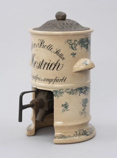 Pojemnik na musztardę - ujęcie z boku; Cylindryczny, porcelitowy pojemnik w kolorze kremowym na wysokiej podstawie z dozownikiem i pokrywą. Stożkowata blaszana pokrywka z wypłaszczonym rondem i okrągłym uchwytem, fakturowana cyzelowanymi wydłużonymi owalami. Pojemnik cylindryczny. Z prawej wklęsły od spodu, zaokrąglony uchwyt. Na górnej części wyśrodkowany, czarny, ozdobny napis pisanką w trzech wierszach, z których pierwszy zapisany po łuku. Porcelitowa podstawa pojemnika w formie pustego cylindra bez dna z wywiniętą na zewnątrz dolną krawędzią markującą stopę oraz plastycznym pierścieniem nieco powyżej. Przed wypałem w podstawie wycięta pionowo część obwodu stanowi prostokątny otwór, z którego wystaje metalowy wąski dozownik. Dekorowany granatową kalkomanią: na podstawie, brzegach obu porcelitowych części wzór rocailliowy w dookolnych pasmach, na uchwytach oraz punktowo na podstawie i pojemniku gałązki róż.