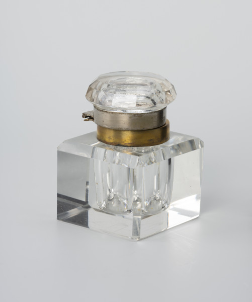 Kałamarz - ujęcie ze skosu z drugiej strony; Kałamarz ze szkła kryształowego z metalowym kołnierzem oprawy ujmującej pokrywę. Pojemnik w formie graniastosłupa z szeroko sfazowanymi krawędziami i wklęsłym cylindrycznym wnętrzem o średnicy 2.25 cm przeznaczonym na atrament. Ponad otworem kołnierz ujęty w metalową oprawę z posrebrzanego mosiądzu. Pokrywa ze szkła lanego z zagłębionym wnętrzem i kołnierzem analogicznym jak w części głównej, ujętym w metalową oprawę z posrebrzanego mosiądzu, która ujmuje kołnierz pierścieniem z otokiem. Górna część pokrywy okrągła z podwójnym szlifem tworzącym 12-ścienne łagodne zwężenie. Część korpusowa dzięki kryształowej przejrzystości tworzy zwierciadlane odbicia, powielające efekt szlifowanych krawędzi naczynia.