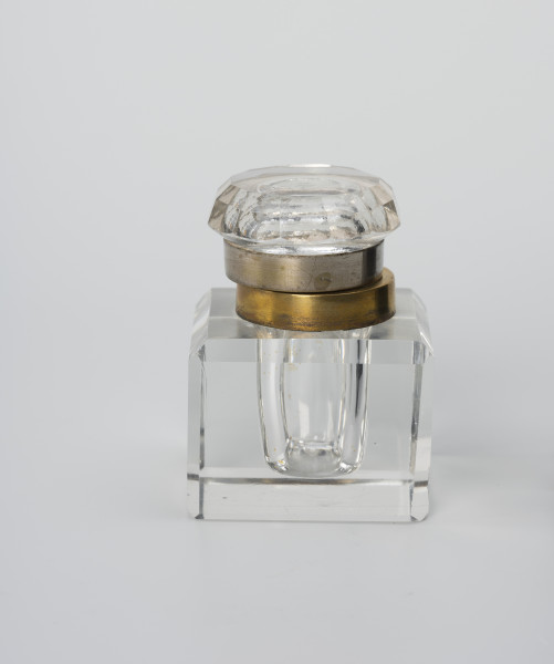 Kałamarz - ujęcie z drugiego boku; Kałamarz ze szkła kryształowego z metalowym kołnierzem oprawy ujmującej pokrywę. Pojemnik w formie graniastosłupa z szeroko sfazowanymi krawędziami i wklęsłym cylindrycznym wnętrzem o średnicy 2.25 cm przeznaczonym na atrament. Ponad otworem kołnierz ujęty w metalową oprawę z posrebrzanego mosiądzu. Pokrywa ze szkła lanego z zagłębionym wnętrzem i kołnierzem analogicznym jak w części głównej, ujętym w metalową oprawę z posrebrzanego mosiądzu, która ujmuje kołnierz pierścieniem z otokiem. Górna część pokrywy okrągła z podwójnym szlifem tworzącym 12-ścienne łagodne zwężenie. Część korpusowa dzięki kryształowej przejrzystości tworzy zwierciadlane odbicia, powielające efekt szlifowanych krawędzi naczynia.