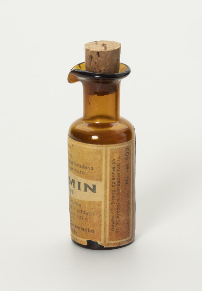 butelka po lekarstwach - ujęcie z prawego boku; Niewielka buteleczka z brązowego szkła. Korpus walcowy, szyjka prosta walcowa przechodząca w wywinięty na zewnątrz kołnierz, w którym wykształcony jest niewielki dziubek. Etykieta w obrębie całego korpusu brązowa z poziomym białym pasem w centrum i napisem czarnymi literami. W białej ramce wiodoczny napis: Dawka 2-3 razy dziennie po 20-30 kropel w herbacie lub tp. Wywóz wzbroniony Nr. rej. 903. Zamknięcie buteleczki korkiem ręcznie krojonym do otworu.