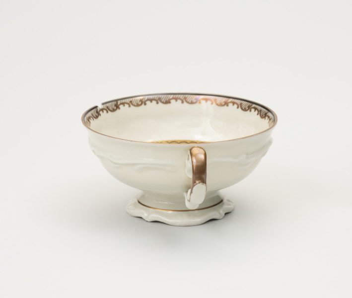 Filiżanka do herbaty - ujęcie z boku; Duża filiżanka z białej porcelany dekorowana złoceniami. Czarka filiżanki okrągła w przekroju poziomym, szeroka na wysokości wylewu zwęża się w kierunku dna. Wewnątrz poniżej wylewu złocony ornament w formie ukośnego szrafowania ograniczonego od góry poziomą dookolną linią i od dołu ułożonymi na przemian C-kształtnymi elementami nawiązującymi do uproszczonego ornamentu małżowinowo-chrząstkowego. Na zewnętrznej ściance czarki oraz na stopce delikatne dookolne pasmo ornamentu plastycznego w formie spłaszczonych esownic i rozciągniętego motywu małżowinowo-chrząstkowego. Nawiązuje do niego widoczna na przodzie forma uszka ozdobionego pasmem złocenia na stronie grzbietowej, które przechodzi w cienką strużkę podkreślającą plastyczność ślimacznicy na profilu uszka, tuż przy łączeniu górnej jego części z czarką.
