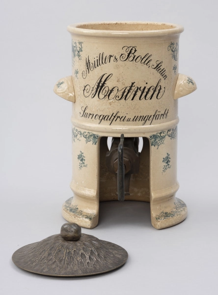 Pojemnik na musztardę - ujęcie z przodu; Cylindryczny, porcelitowy pojemnik w kolorze kremowym na wysokiej podstawie z dozownikiem. Przed nim leży do niego pokrywa, stożkowata blaszana z wypłaszczonym rondem i okrągłym uchwytem, fakturowana cyzelowanymi wydłużonymi owalami. Pojemnik cylindryczny z dwoma wklęsłymi od spodu, zaokrąglonymi uchwytami, poziomo po bokach. Na górnej części wyśrodkowany, czarny, ozdobny napis pisany w trzech wierszach, z których pierwszy zapisany po łuku. Porcelitowa podstawa pojemnika w formie pustego cylindra bez dna z wywiniętą na zewnątrz dolną krawędzią markującą stopę oraz plastycznym pierścieniem nieco powyżej. Przed wypałem w podstawie wycięta pionowo część obwodu stanowi prostokątny otwór, z którego wystaje metalowy wąski dozownik. Dekorowany granatową kalkomanią: na podstawie, brzegach obu porcelitowych części wzór rocailliowy w dookolnych pasmach, na uchwytach oraz punktowo na podstawie i pojemniku gałązki róż.