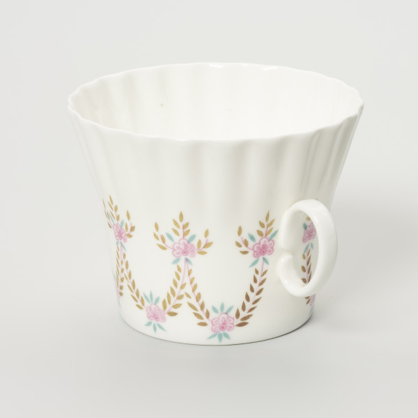 Filiżanka - ujęcie z boku; Duża filiżanka z białej porcelany o lekko dzwonowatej formie, z płaskim dnem i karbowaną górną częścią czarki. Uszko proste, owalne doklejone pośrodku wysokości. Dekorowana malowanymi festonami z wici roślinno-kwiatowej w barwach różu, zieleni i starego złota.
