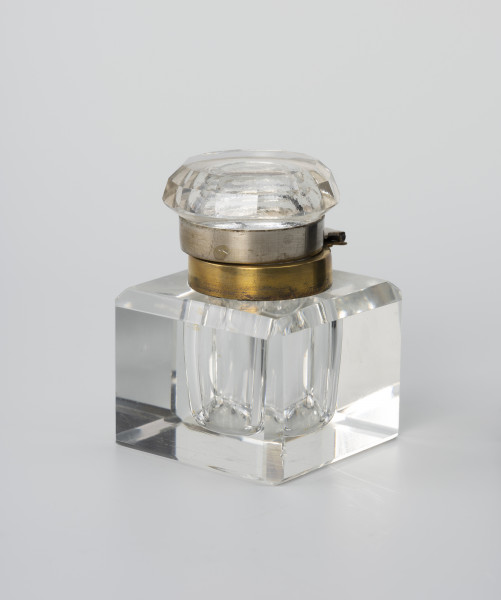 Kałamarz - ujęcie ze skosu; Kałamarz ze szkła kryształowego z metalowym kołnierzem oprawy ujmującej pokrywę. Pojemnik w formie graniastosłupa z szeroko sfazowanymi krawędziami i wklęsłym cylindrycznym wnętrzem o średnicy 2.25 cm przeznaczonym na atrament. Ponad otworem kołnierz ujęty w metalową oprawę z posrebrzanego mosiądzu. Pokrywa ze szkła lanego z zagłębionym wnętrzem i kołnierzem analogicznym jak w części głównej, ujętym w metalową oprawę z posrebrzanego mosiądzu, która ujmuje kołnierz pierścieniem z otokiem. Górna część pokrywy okrągła z podwójnym szlifem tworzącym 12-ścienne łagodne zwężenie. Część korpusowa dzięki kryształowej przejrzystości tworzy zwierciadlane odbicia, powielające efekt szlifowanych krawędzi naczynia.