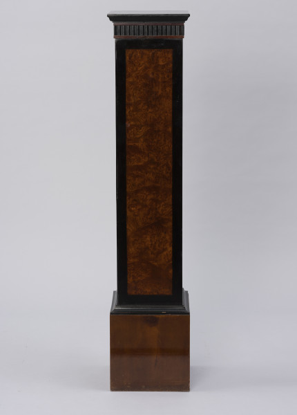 Postument dekoracyjny - ujęcie prosto; Wolnostojący wysoki postument drewniany, fornirowany, z profilowaną krawędzią płyty wieńczącej i poszerzonym cokołem wykończonym profilowaną listwą. Pod płytą wieńczącą fryz z ciemnego, czarnego drewna z dekoracją w formie wąskich pionowych regularnych wyżłobień. Dzięki zastosowaniu różnych gatunków i barw forniru płyciny pionowych ścianek postumentu uzyskały ciemne, czarne obramienie w formie pasa przy krawędziach. Na płyciny ścianek pomiędzy obramieniem wykorzystano jaśniejsze miodowe drewno o bogatym naturalnym wzorze czeczotowym.