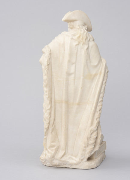 Miniatura szczecińskiego pomnika króla Fryderyka II Wielkiego - ujęcie z tyłu; Alabastrowa rzeźba przedstawiająca stojącą tyłem postać. Postać z głową zwróconą lekko w lewą stronę. Na głowie trójgraniasty kapelusz, spod którego wystają krótkie lokowane pukle włosów, na środku spięte włosy w dłuższą kitkę. Na plecach długi futrzany płaszcz.