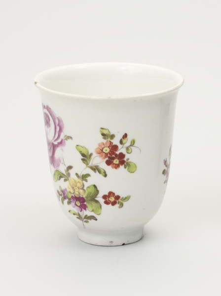 Czarka - ujęcie z przodu; Smukła czarka na stopie o dzwonowatej formie z białej porcelany udekorowana malowanymi różnobarwnymi piwoniami. Na pędzie głównym drobne kwiaty w kolorze czerwonym, żółtym i różowym oraz gałązki z zielonymi listkami. Na lewym pędzie fragment dużego, pełnego, rozwiniętego kwiata.