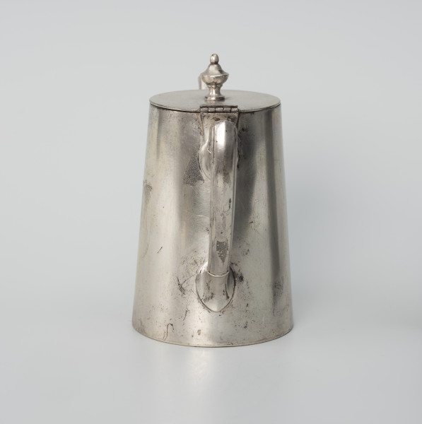 Dzbanuszek z zastawy Konzerthaus Stettin - ujęcie ze skosu z prawej; Dzbanuszek o prostej, zgeometryzowanej formie, bez wykształconej stopy. Korpus cylindryczny, zwężający się ku górze, zamykany płaską, okrągłą pokrywą z gałką w centrum. Ucho o czworobocznym przekroju, górą wyniesione do wysokości pokrywy, a następnie załamane, poprowadzone niemal poziomo i powtórnie załamane, a dalej delikatnie wygięte i łukiem wprowadzone do ścianki korpusu. Górą i dołem powyżej wygięcia przewiązka. Pokrywa - krążek z blachy na zawiasie powyżej ucha; uchwyt umocowany centralnie, w formie kielichowej gałki zwieńczonej kulką.