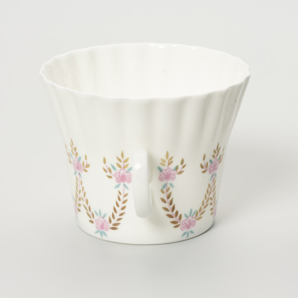 Filiżanka - ujęcie z boku; Duża filiżanka z białej porcelany o lekko dzwonowatej formie, z płaskim dnem i karbowaną górną częścią czarki. Uszko na wprost, owalne doklejone pośrodku wysokości. Dekorowana malowanymi festonami z wici roślinno-kwiatowej w barwach różu, zieleni i starego złota.