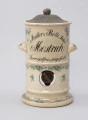 Pojemnik na musztardę - ujęcie z tyłu; Cylindryczny, porcelitowy pojemnik w kolorze kremowym na wysokiej podstawie z dozownikiem i pokrywą. Stożkowata blaszana pokrywka z wypłaszczonym rondem i okrągłym uchwytem, fakturowana cyzelowanymi wydłużonymi owalami. Pojemnik cylindryczny z dwoma wklęsłymi od spodu, zaokrąglonymi uchwytami, poziomo po bokach. Na górnej części wyśrodkowany, czarny, ozdobny napis pisanką w trzech wierszach, z których pierwszy zapisany po łuku. Porcelitowa podstawa pojemnika z wywiniętą na zewnątrz dolną krawędzią markującą stopę oraz plastycznym pierścieniem nieco powyżej, nad nim okrągły otwór z widocznym metalowym pokrętłem. Pojemnik dekorowany granatową kalkomanią: na podstawie, brzegach obu porcelitowych części wzór rocailliowy w dookolnych pasmach, na uchwytach oraz punktowo na podstawie i pojemniku gałązki róż.
