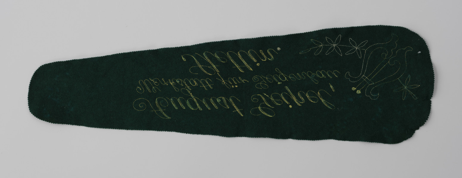 sukno z futerału skrzypiec - ujęcie odwrocia haftowanego sukna; Sukno ochronne z zielonego aksamitu z wyposażenia futerału skrzypiec z haftowanym żółtą nicią znakiem firmowym: lira z ornamentem kwiatowym z prawej oraz napisem w 3 wierszach. Lira prosta, schematyczna, ułożona ukośnie w lewą stronę. Wyhaftowane kontury oraz ornament kwiatowy wychodzący z obu boków liry i wygięty w kształt łuku. Napis kursywą, ozdobną czcionką, ręcznie haftowaną. Brzegi sukna ząbkowane.