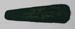 sukno z futerału skrzypiec - ujęcie odwrocia haftowanego sukna; Sukno ochronne z zielonego aksamitu z wyposażenia futerału skrzypiec z haftowanym żółtą nicią znakiem firmowym: lira z ornamentem kwiatowym z prawej oraz napisem w 3 wierszach. Lira prosta, schematyczna, ułożona ukośnie w lewą stronę. Wyhaftowane kontury oraz ornament kwiatowy wychodzący z obu boków liry i wygięty w kształt łuku. Napis kursywą, ozdobną czcionką, ręcznie haftowaną. Brzegi sukna ząbkowane.
