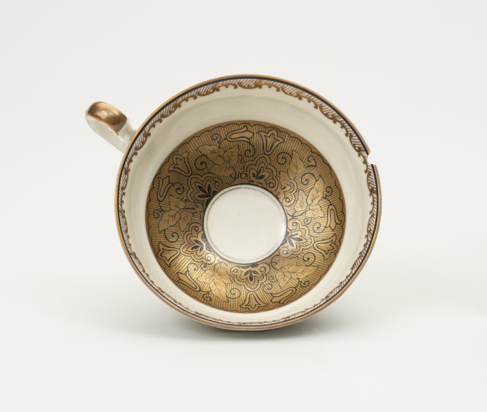 Filiżanka do herbaty - ujęcie z góry; Duża filiżanka z białej porcelany dekorowana złoceniami. Czarka filiżanki okrągła w przekroju poziomym. Wewnątrz w dolnej części czarki szeroki złocony pas z konturowym ornamentem roślinnym, a poniżej wylewu złocony ornament w formie ukośnego szrafowania ograniczonego od góry poziomą dookolną linią i od dołu ułożonymi na przemian C-kształtnymi elementami nawiązującymi do uproszczonego ornamentu małżowinowo-chrząstkowego. Widoczne uszko ozdobione pasmem złocenia na stronie grzbietowej.