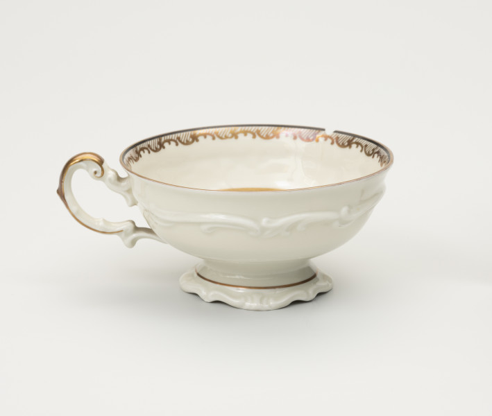 Filiżanka do herbaty - ujęcie z tyłu; Duża filiżanka z białej porcelany dekorowana złoceniami. Czarka filiżanki okrągła w przekroju poziomym, szeroka na wysokości wylewu zwęża się w kierunku dna. Wewnątrz złocony ornament w formie ukośnego szrafowania ograniczonego od góry poziomą dookolną linią i od dołu ułożonymi na przemian C-kształtnymi elementami nawiązującymi do uproszczonego ornamentu małżowinowo-chrząstkowego. Na zewnętrznej ściance czarki oraz na stopce delikatne dookolne pasmo ornamentu plastycznego w formie spłaszczonych esownic i rozciągniętego motywu małżowinowo-chrząstkowego. Nawiązuje do niego również forma uszka z lewej strony ozdobionego pasmem złocenia na stronie grzbietowej, które przechodzi w cienką strużkę podkreślającą plastyczność ślimacznicy na profilu uszka, tuż przy łączeniu górnej jego części z czarką.