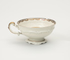 Filiżanka do herbaty - ujęcie z tyłu; Duża filiżanka z białej porcelany dekorowana złoceniami. Czarka filiżanki okrągła w przekroju poziomym, szeroka na wysokości wylewu zwęża się w kierunku dna. Wewnątrz złocony ornament w formie ukośnego szrafowania ograniczonego od góry poziomą dookolną linią i od dołu ułożonymi na przemian C-kształtnymi elementami nawiązującymi do uproszczonego ornamentu małżowinowo-chrząstkowego. Na zewnętrznej ściance czarki oraz na stopce delikatne dookolne pasmo ornamentu plastycznego w formie spłaszczonych esownic i rozciągniętego motywu małżowinowo-chrząstkowego. Nawiązuje do niego również forma uszka z lewej strony ozdobionego pasmem złocenia na stronie grzbietowej, które przechodzi w cienką strużkę podkreślającą plastyczność ślimacznicy na profilu uszka, tuż przy łączeniu górnej jego części z czarką.