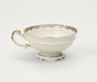 Filiżanka do herbaty - ujęcie z tyłu; Duża filiżanka z białej porcelany dekorowana złoceniami. Czarka filiżanki okrągła w przekroju poziomym, szeroka na wysokości wylewu zwęża się w kierunku dna. Wewnątrz złocony ornament w formie ukośnego szrafowania ograniczonego od góry poziomą dookolną linią i od dołu ułożonymi na przemian C-kształtnymi elementami nawiązującymi do uproszczonego ornamentu małżowinowo-chrząstkowego. Na zewnętrznej ściance czarki oraz na stopce delikatne dookolne pasmo ornamentu plastycznego w formie spłaszczonych esownic i rozciągniętego motywu małżowinowo-chrząstkowego. Nawiązuje do niego również forma uszka z lewej strony ozdobionego pasmem złocenia na stronie grzbietowej, które przechodzi w cienką strużkę podkreślającą plastyczność ślimacznicy na profilu uszka, tuż przy łączeniu górnej jego części z czarką.