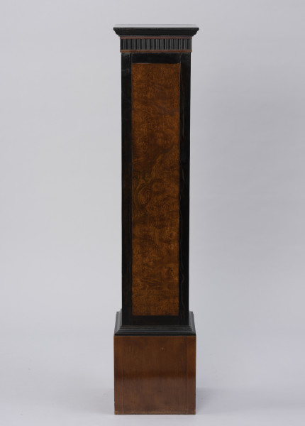 Postument dekoracyjny - ujęcie z boku; Wolnostojący wysoki postument drewniany, fornirowany, z profilowaną krawędzią płyty wieńczącej i poszerzonym cokołem wykończonym profilowaną listwą. Pod płytą wieńczącą fryz z ciemnego, czarnego drewna z dekoracją w formie wąskich pionowych regularnych wyżłobień. Dzięki zastosowaniu różnych gatunków i barw forniru płyciny pionowych ścianek postumentu uzyskały ciemne, czarne obramienie w formie pasa przy krawędziach. Na płyciny ścianek bocznych wykorzystano jaśniejsze miodowe drewno o bogatym naturalnym wzorze czeczotowym.