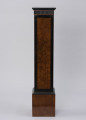 Postument dekoracyjny - ujęcie z boku; Wolnostojący wysoki postument drewniany, fornirowany, z profilowaną krawędzią płyty wieńczącej i poszerzonym cokołem wykończonym profilowaną listwą. Pod płytą wieńczącą fryz z ciemnego, czarnego drewna z dekoracją w formie wąskich pionowych regularnych wyżłobień. Dzięki zastosowaniu różnych gatunków i barw forniru płyciny pionowych ścianek postumentu uzyskały ciemne, czarne obramienie w formie pasa przy krawędziach. Na płyciny ścianek bocznych wykorzystano jaśniejsze miodowe drewno o bogatym naturalnym wzorze czeczotowym.