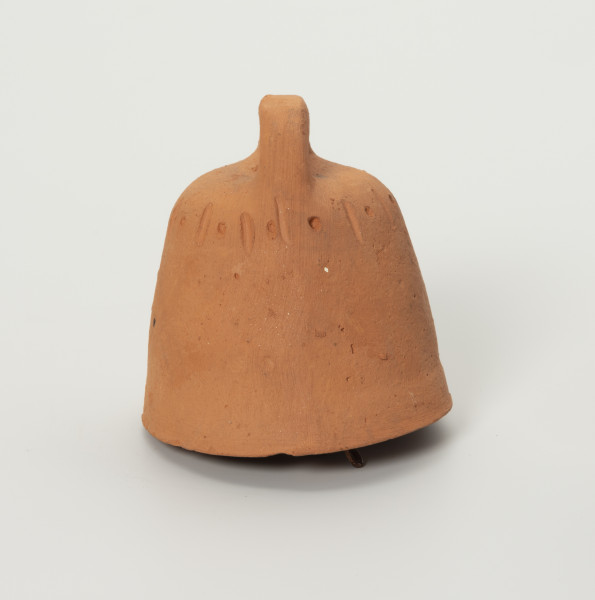 Dzwonek Heleny Kurcyusz - ujęcie z boku; Gliniany dzwonek kloszowy, bez wyraźnie wyodrębnionego płaszcza i wieńca. Szeroka korona w dwóch przeciwległych miejscach nachodzi na brzegi hełmu i górnej krawędzi. Zdobiony przy krawędzi górnej prostym, dookolnym ornamentem z rytych naprzemiennie pionowych kresek i punktów.