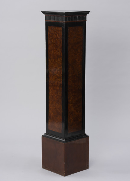 Postument dekoracyjny - ujęcie ze skosu; Wolnostojący wysoki postument drewniany, fornirowany, z profilowaną krawędzią płyty wieńczącej i poszerzonym cokołem wykończonym profilowaną listwą. Pod płytą wieńczącą fryz z ciemnego, czarnego drewna z dekoracją w formie wąskich pionowych regularnych wyżłobień. Dzięki zastosowaniu różnych gatunków i barw forniru płyciny pionowych ścianek postumentu uzyskały ciemne, czarne obramienie w formie pasa przy krawędziach. Na płyciny ścianek bocznych wykorzystano jaśniejsze miodowe drewno o bogatym naturalnym wzorze czeczotowym.