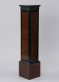 Postument dekoracyjny - ujęcie ze skosu; Wolnostojący wysoki postument drewniany, fornirowany, z profilowaną krawędzią płyty wieńczącej i poszerzonym cokołem wykończonym profilowaną listwą. Pod płytą wieńczącą fryz z ciemnego, czarnego drewna z dekoracją w formie wąskich pionowych regularnych wyżłobień. Dzięki zastosowaniu różnych gatunków i barw forniru płyciny pionowych ścianek postumentu uzyskały ciemne, czarne obramienie w formie pasa przy krawędziach. Na płyciny ścianek bocznych wykorzystano jaśniejsze miodowe drewno o bogatym naturalnym wzorze czeczotowym.