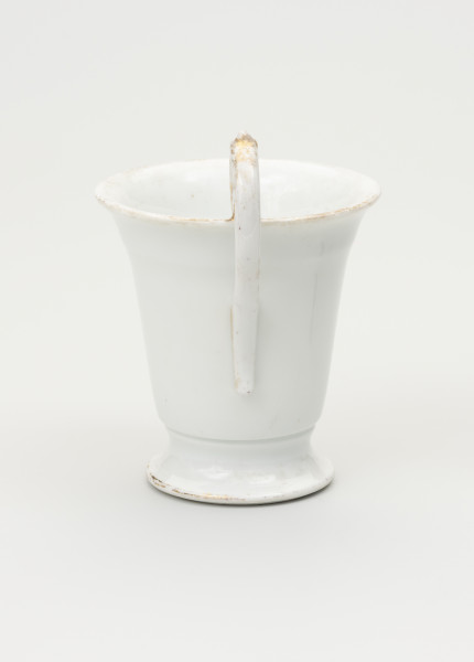 Filiżanka - ujęcie z tyłu; Filiżanka z grubej, białej porcelany w kształcie odwróconego dzwonu. Czara wysoka, łagodnie rozszerzająca się ku górze, z wylewem wywiniętym na zewnątrz. Stopa niska, okrągła, rozpłaszczona na zewnątrz, z pierścieniem na styku z czarą. Uszko wysokie, wyniesione ponad poziom wylewu, doklejone do ścianki czary i wnętrza jej wylewu.