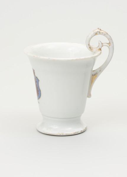 Filiżanka - ujęcie z boku; Filiżanka z grubej, białej porcelany w kształcie odwróconego dzwonu. Czara wysoka, łagodnie rozszerzająca się ku górze, z wylewem wywiniętym na zewnątrz. Stopa niska, okrągła, rozpłaszczona na zewnątrz, z pierścieniem na styku z czarą. Uszko wysokie, wyniesione ponad poziom wylewu w kształcie zwiniętego zwieńczenia pastorału; doklejone do ścianki czary i wnętrza jej wylewu. Uszko posiada widoczne przetarte złocenia. Czara ozdobiona w centrum, po przeciwnej stronie uszka. Ślady złoceń wokół wylewu od środka czary, na brzegu stopy oraz na uchwycie.