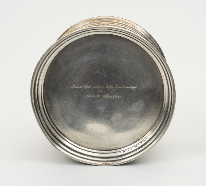 Puszka liturgiczna na komunikanty - ujęcie z dołu; Okrągła puszka liturgiczna na komunikanty, wykonana z pozłacanego srebra. Od spodu, na środku napis ryty kursywą w dwóch liniach: Friedr. Ebel entn. Wilh. Arnolt (...)gef. | 29.6.99.Berlin. Pomiędzy linijkami wybite wklęsłe punce: ptak, zamknięta korona, cyfra 80C oraz ostatnia niezidentyfikowana punca.