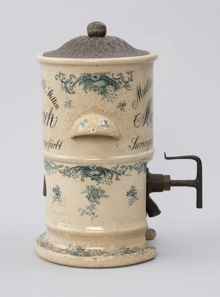Pojemnik na musztardę - ujęcie z lewego boku; Cylindryczny, porcelitowy pojemnik w kolorze kremowym na wysokiej podstawie z dozownikiem i pokrywą. Stożkowata blaszana pokrywka z wypłaszczonym rondem i okrągłym uchwytem, fakturowana cyzelowanymi wydłużonymi owalami. Pojemnik cylindryczny z wystającym z przodu wklęsłymi od spodu, zaokrąglonym uchwytem. Po bokach fragmenty w górnej części wyśrodkowane, czarne, ozdobne napisy pisane w trzech wierszach, z których pierwszy zapisany po łuku. Porcelitowa podstawa pojemnika w formie pustego cylindra bez dna z wywiniętą na zewnątrz dolną krawędzią markującą stopę oraz plastycznym pierścieniem nieco powyżej. Z prawej strony wystaje metalowy wąski dozownik. Pojemnik dekorowany granatową kalkomanią: na podstawie, brzegach obu porcelitowych części wzór rocailliowy w dookolnych pasmach, na uchwytach oraz punktowo na podstawie i pojemniku gałązki róż.
