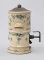 Pojemnik na musztardę - ujęcie z lewego boku; Cylindryczny, porcelitowy pojemnik w kolorze kremowym na wysokiej podstawie z dozownikiem i pokrywą. Stożkowata blaszana pokrywka z wypłaszczonym rondem i okrągłym uchwytem, fakturowana cyzelowanymi wydłużonymi owalami. Pojemnik cylindryczny z wystającym z przodu wklęsłymi od spodu, zaokrąglonym uchwytem. Po bokach fragmenty w górnej części wyśrodkowane, czarne, ozdobne napisy pisane w trzech wierszach, z których pierwszy zapisany po łuku. Porcelitowa podstawa pojemnika w formie pustego cylindra bez dna z wywiniętą na zewnątrz dolną krawędzią markującą stopę oraz plastycznym pierścieniem nieco powyżej. Z prawej strony wystaje metalowy wąski dozownik. Pojemnik dekorowany granatową kalkomanią: na podstawie, brzegach obu porcelitowych części wzór rocailliowy w dookolnych pasmach, na uchwytach oraz punktowo na podstawie i pojemniku gałązki róż.