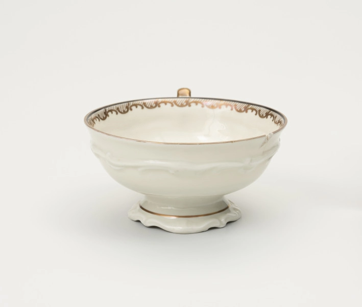 Filiżanka do herbaty - ujęcie z drugiego boku; Duża filiżanka z białej porcelany dekorowana złoceniami. Czarka filiżanki okrągła w przekroju poziomym, szeroka na wysokości wylewu zwęża się w kierunku dna. Wewnątrz poniżej wylewu złocony ornament w formie ukośnego szrafowania ograniczonego od góry poziomą dookolną linią i od dołu ułożonymi na przemian C-kształtnymi elementami nawiązującymi do uproszczonego ornamentu małżowinowo-chrząstkowego. Na zewnętrznej ściance czarki oraz na stopce delikatne dookolne pasmo ornamentu plastycznego w formie spłaszczonych esownic i rozciągniętego motywu małżowinowo-chrząstkowego.