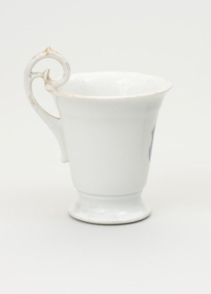 Filiżanka - ujęcie z drugiego boku; Filiżanka z grubej, białej porcelany w kształcie odwróconego dzwonu. Czara wysoka, łagodnie rozszerzająca się ku górze, z wylewem wywiniętym na zewnątrz. Stopa niska, okrągła, rozpłaszczona na zewnątrz, z pierścieniem na styku z czarą. Uszko wysokie, wyniesione ponad poziom wylewu w kształcie zwiniętego zwieńczenia pastorału; doklejone do ścianki czary i wnętrza jej wylewu. Ślady złoceń wokół wylewu od środka czary, na brzegu stopy oraz na uchwycie.