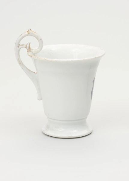 Filiżanka - ujęcie z drugiego boku; Filiżanka z grubej, białej porcelany w kształcie odwróconego dzwonu. Czara wysoka, łagodnie rozszerzająca się ku górze, z wylewem wywiniętym na zewnątrz. Stopa niska, okrągła, rozpłaszczona na zewnątrz, z pierścieniem na styku z czarą. Uszko wysokie, wyniesione ponad poziom wylewu w kształcie zwiniętego zwieńczenia pastorału; doklejone do ścianki czary i wnętrza jej wylewu. Ślady złoceń wokół wylewu od środka czary, na brzegu stopy oraz na uchwycie.