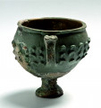 Zrekonstruowany ceramiczny pucharek na cylindrycznej nóżce zakończonej kolistą stopką, ukazany z boku od strony uszka. Wyraźnie widoczne gładsze obszary zrekonstruowane i mocno chropowate po przepaleniu elementy oryginalne.