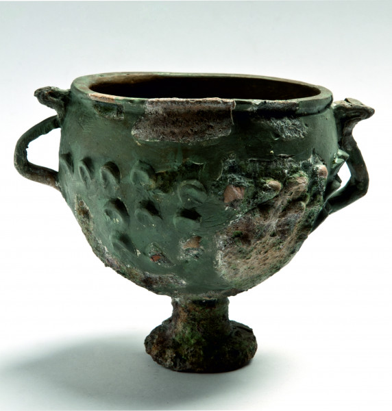 Zrekonstruowany ceramiczny pucharek na cylindrycznej nóżce zakończonej kolistą stopką, ukazany od frontu. Po bokach widoczne esowate uszka spłaszczone od góry. Wyraźnie widoczne gładsze obszary zrekonstruowane i mocno chropowate po przepaleniu elementy oryginalne.
