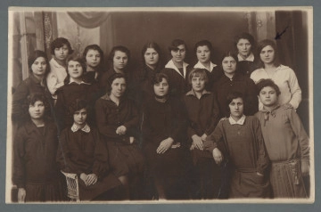 MPOLIN-A48.1.2 Fotografia osiemnastu dziewcząt, ubranych elegancko - w białe koszule i kostiumy czy sukienki wyjściowe - w trzech rzędach (w najniższym siedzą i klęczą); jak możemy się domyślać (stąd, że nazwiska powtarzają się we wpisach pamiętnikowych Mani Żelaznej z archiwaliów przekazanych do Korczakianum), są to wychowanki internatu: Głównego Domu Schronienia w Warszawie przy ul. Wolskiej 18. Wśród nich Mania Żelazna, najbardziej po prawej u góry, oznaczona strzałką po prawej. W górnej części widoczne atelierowe tło - być może przedstawienie wnętrza pałacu (?). Fotografia z białym wąskim marginesem wzdłuż prostych krawędzi. W prawym dolnym rogu rewersu zamazanie nieznanej inskrypcji napisanej czarnym piórem - zamazanie niebieskim długopisem.