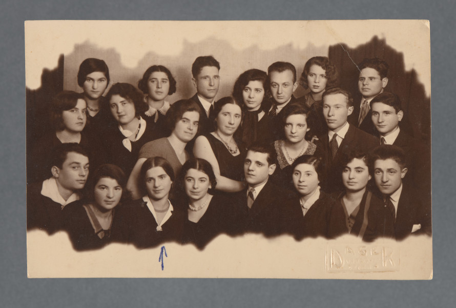 Fotografia grupowa, ustawiana, jaw wynika z opisu - wykonana z okazji wyjazdu koleżanki Mani Żelaznej do Palestyny - Sary, być może Sary Tenenbaum, widocznej, jak się wydaje, w dolnym rzędzie (trzecia od prawej?). Pośrodku przedstawienia wychowawczyni? Znana z innych zdjęć kolekcji, jak się wydaje - m.in. 1.16, 1.28.Mani Żelazna wskazana znakiem długopisowym - strzałką na dolnym marginesie. Wzdłuż dolnej i górnej krawędzi - margines biały o falistej linii, stylizowanej na kształt chmury. Krawędzie odbitki proste.