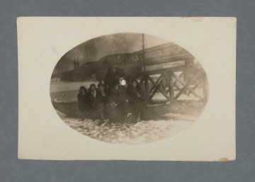 Fotografia monochromatyczna, widocznych sześć kobiet i jeden mężczyzna, przed nimi widoczny śnieg; zdjęcie nieco rozmazane, mocno ciemne, z wadą wywołania pośrodku (biała plamka). W tle most Kierbedzia z charakterystycznymi dwoma zaokrąglonymi przęsłami, widoczne także, po lewej, wieże katedry praskiej (kościół św. Michała Archanioła i św. Floriana Męczennika). Fotografia owalna, na prostokątnym papierze – z białym obramowaniem obrzeża.