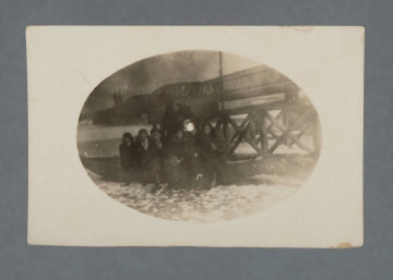Fotografia monochromatyczna, widocznych sześć kobiet i jeden mężczyzna, przed nimi widoczny śnieg; zdjęcie nieco rozmazane, mocno ciemne, z wadą wywołania pośrodku (biała plamka). W tle most Kierbedzia z charakterystycznymi dwoma zaokrąglonymi przęsłami, widoczne także, po lewej, wieże katedry praskiej (kościół św. Michała Archanioła i św. Floriana Męczennika). Fotografia owalna, na prostokątnym papierze – z białym obramowaniem obrzeża.