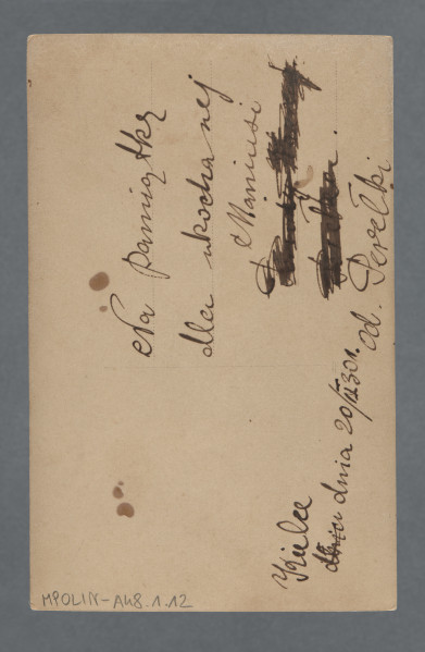 Grupa osób zgromadzonych przed ceglanym nieotynkowanym budynkiem oraz stojących bądź siedzących na desce rozpostartej na konstrukcji balkonowej; budynek za jasnym drewnianym płotem. Sepia.Wśród zgromadzonych znajduje się zapewne osoba pisząca dedykację na odwrocie: Perełka Ajzówna, koleżanka Mani Żelaznej z domu opiekuńczego w Warszawie. Fotografia z prostymi krawędziami, z czterema plamami-przebarwieniami w prawej części, a także przebarwieniem wzdłuż górnej krawędzi.