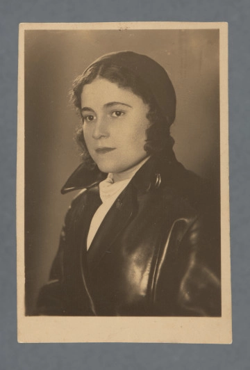 Fotografia monochromatyczna kobiety w półprofilu, popiersie. Włosy z przedziałkiem, za ucho, lokowane, ubrany beret oraz gruba skórzana czarna kurtka, pod nią biała bluzka (?).Na krawędzi dłuższej przy lewym dolnym rogu rewersu ślad po fioletowym tuszu pieczęci (?).Krawędzie odbitki proste.